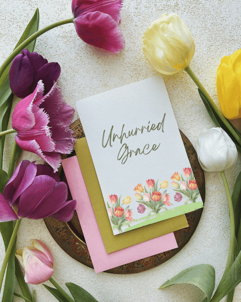 Flatlay of a tulip wreath in soft spring colors encircling a blank cardstock with handwritten text “Unhurried Grace” in olive green, paired with an olive envelope and floral border — symbolizing slow, intentional living with faith and grace.