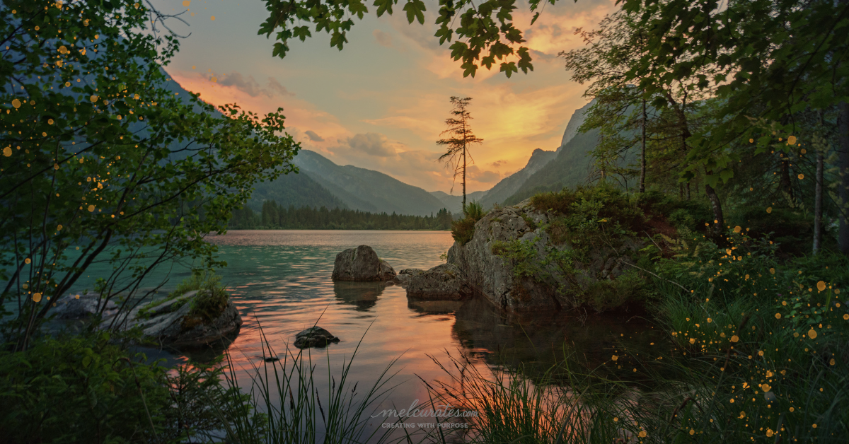 Nature scene at golden hour featuring a blush and orange sky over distant hills and mountains. In the foreground, flowering tree branches frame a shimmering lake with sunlight reflecting off the water. perfect summer gift from God.