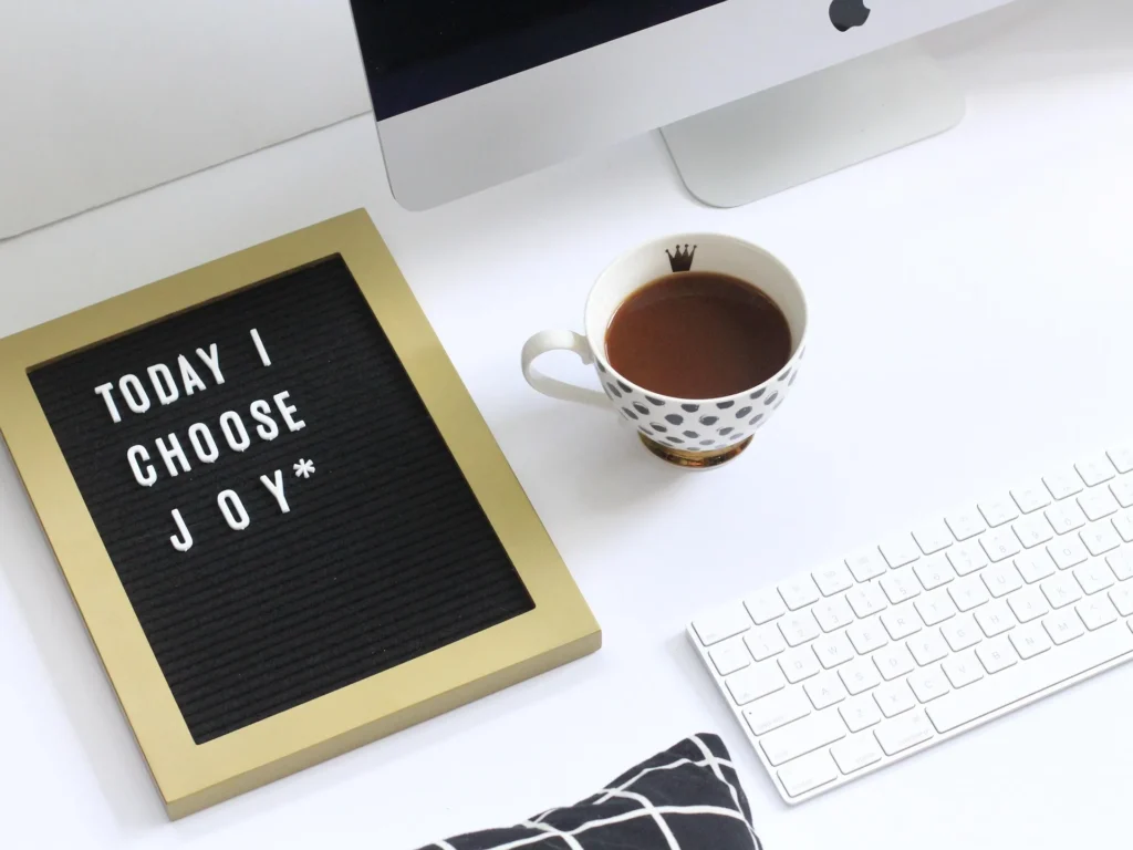 Creative workspace with white keyboard, monitor, cup of tea, and a sign that says ‘Today I Choose Joy’ — a peaceful, faith-inspired setting for connection and encouragement. Contact Me.